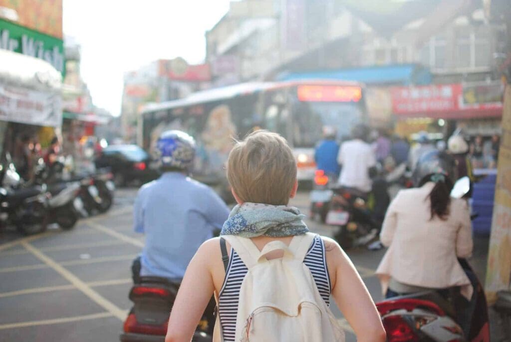 A person with short hair, wearing a striped shirt and a backpack, stands in a crowded street lined with shops. Motorcycles and people move around, and a bus is visible in the background. Urban hustle and bustle surrounds them.