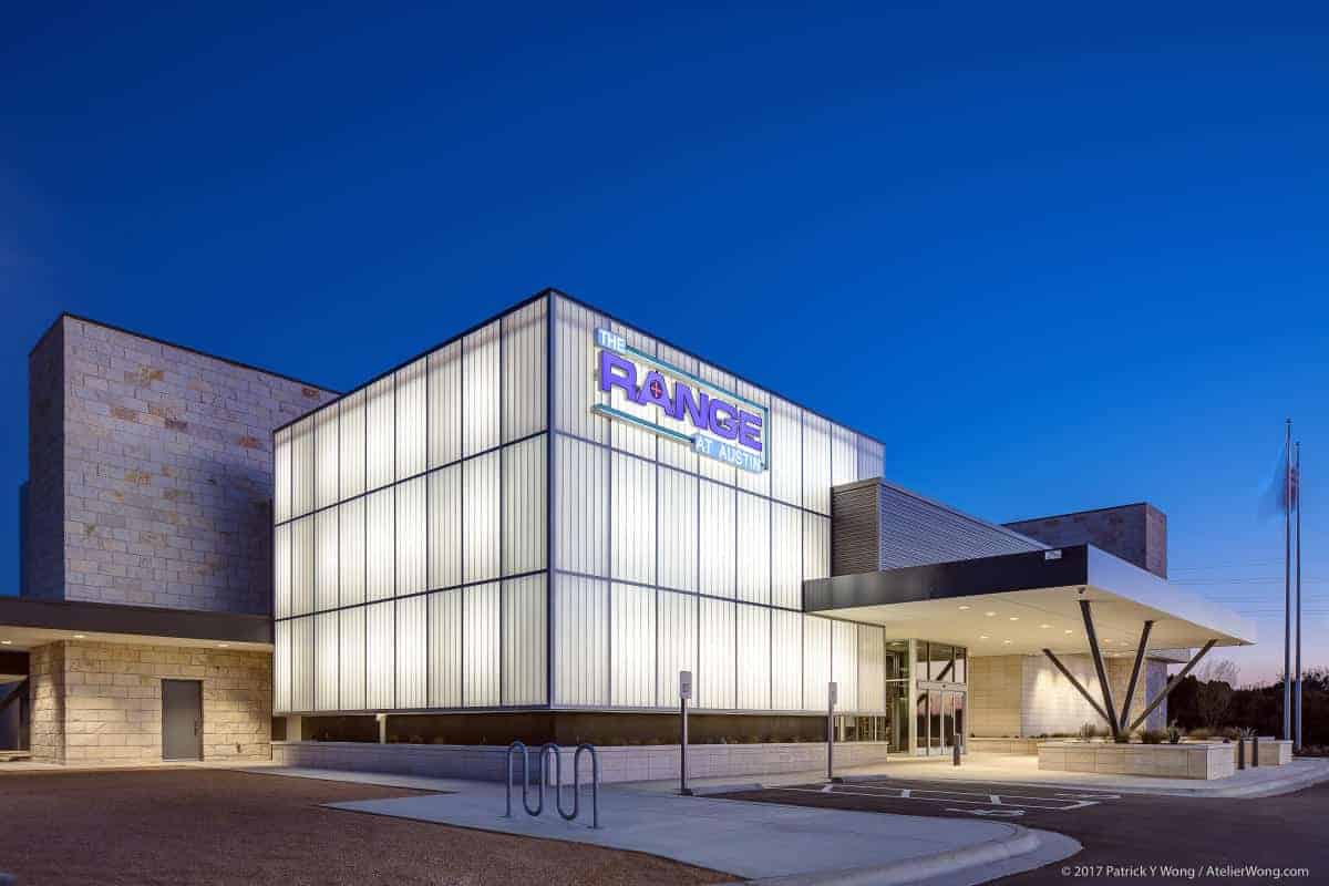 Modern building at dusk with a well-lit, translucent facade and a sign reading RANGE in purple and blue. The structure features angular lines and a flat roof, with a bicycle rack in front and a clear sky in the background.