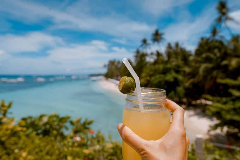 A hand holds a mason jar filled with a yellow drink and a straw, garnished with a slice of lime. In the background, a tropical beach with blue water, white sand, and palm trees is visible under a bright blue sky.