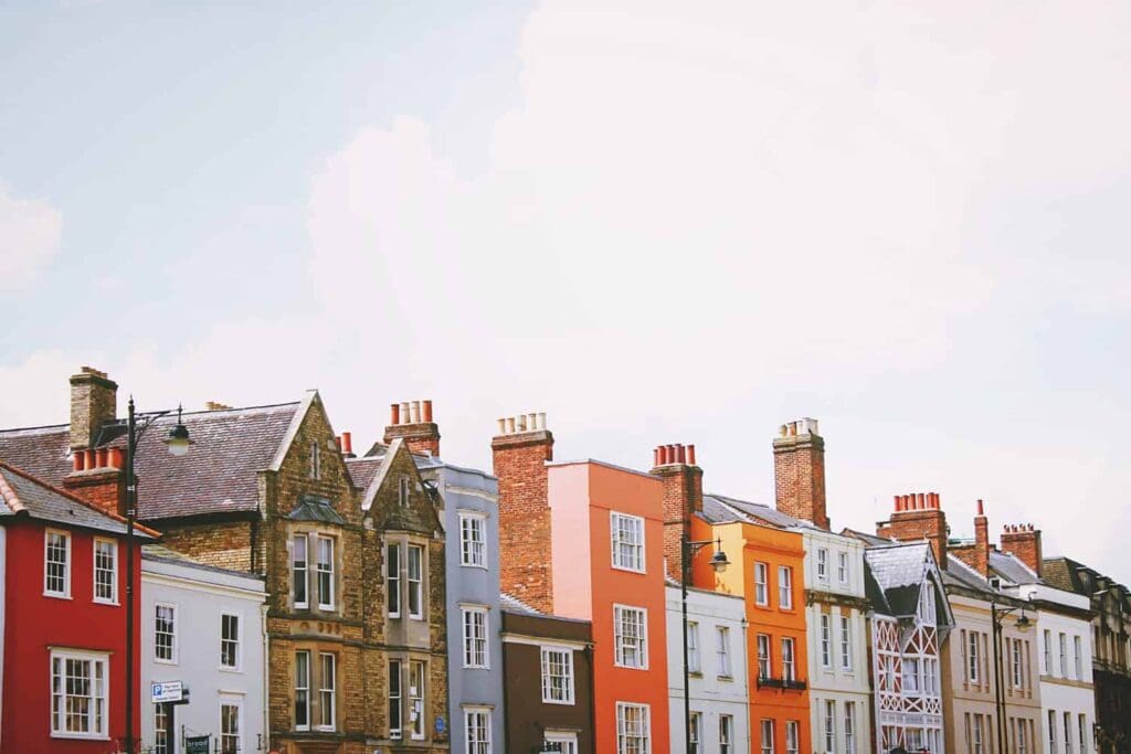 Colorful row of traditional European houses with various architectural styles and chimney stacks against a clear sky. The buildings are painted in red, white, gray, orange, and beige, creating a vibrant streetscape.