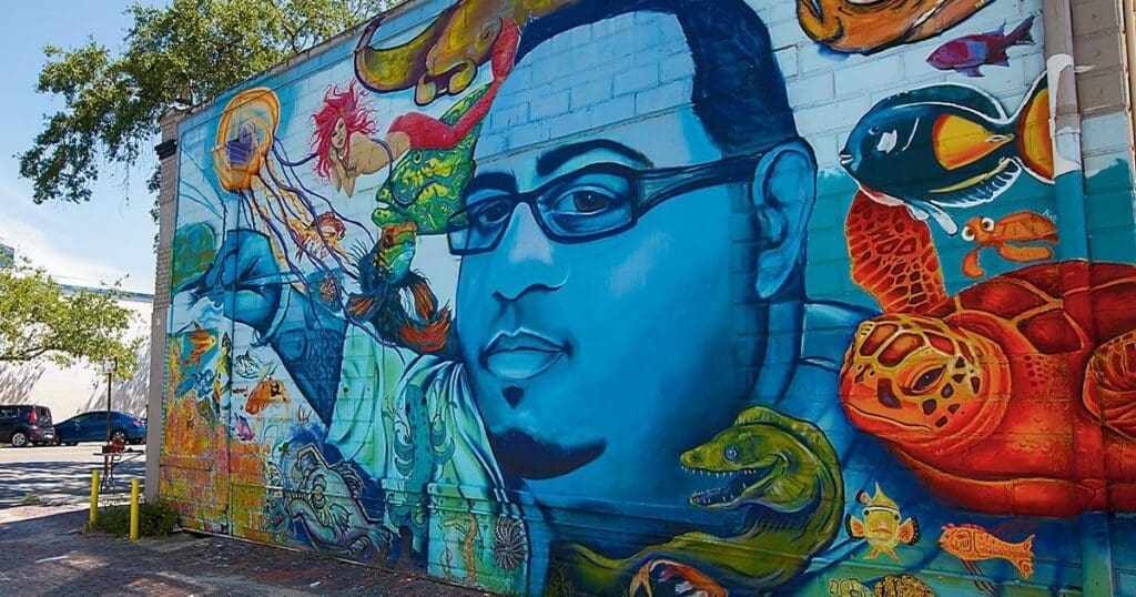 A vibrant mural on a brick wall features a blue-toned portrait of a person with glasses surrounded by colorful marine life, including fish, turtles, and corals. The scene is set in an outdoor urban environment with trees and pavement.