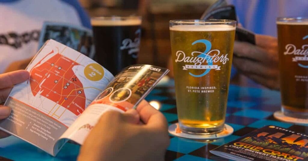 Two people sit at a table with checkered tablecloth, each holding a pint of beer. One person is looking at a colorful booklet showing a map, with another booklet nearby. The beer glasses have 3 Daughters Brewing logos on them.