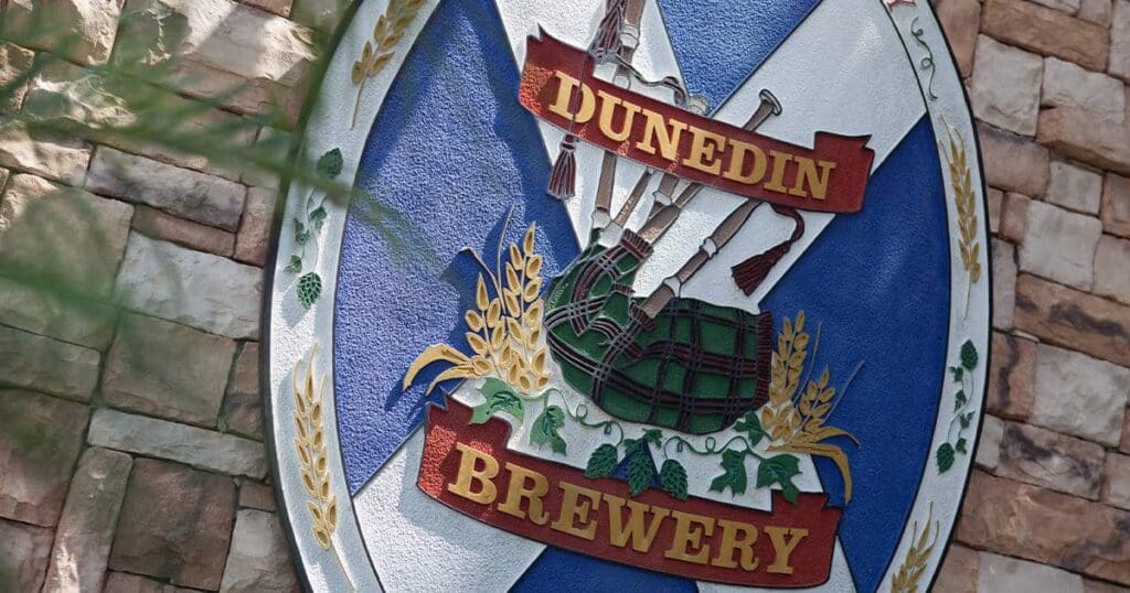 The image shows a circular sign for Dunedin Brewery. It features a Scottish theme with bagpipes, a blue background, and decorative wheat stalks. The sign is mounted on a stone wall.