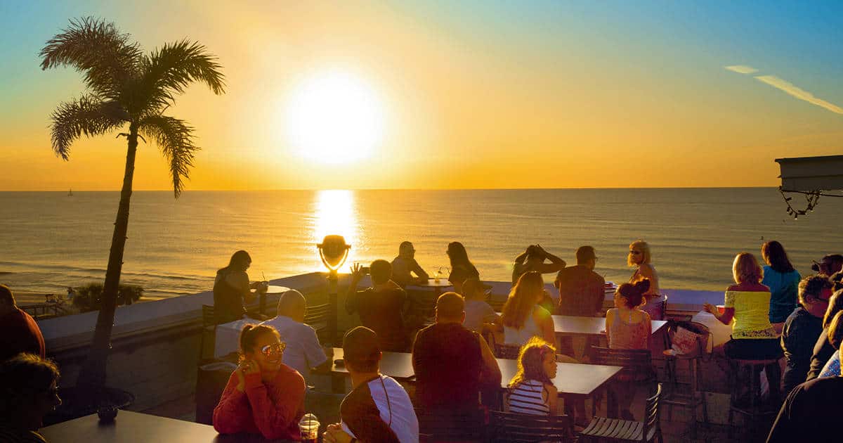 A group of people sitting at tables on a rooftop, enjoying drinks and socializing while watching a vibrant ocean sunset. A palm tree is silhouetted against the sky, and the golden light reflects off the water.