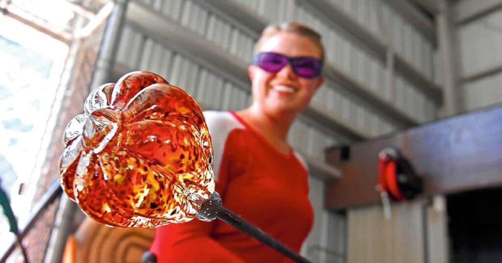 A person wearing purple safety glasses works with molten glass, shaped like a flower, on a rod. They are smiling and wearing a red shirt, with a workshop setting in the background.