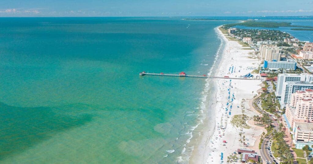 Aerial view of a coastal city with a long pier extending into the ocean. The beach is lined with umbrellas and people, while high-rise buildings and greenery border the shoreline. The water is a mix of turquoise and deep blue.