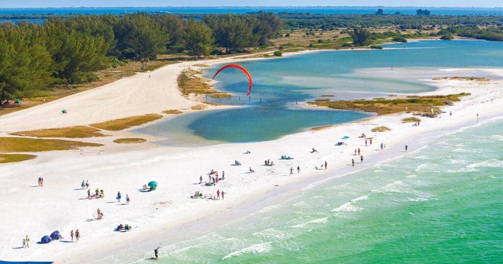 Aerial view of a beach with white sand and greenish-blue water. People are sunbathing and swimming. Trees and vegetation line the shoreline. A red parasail is visible above the water in the distance.