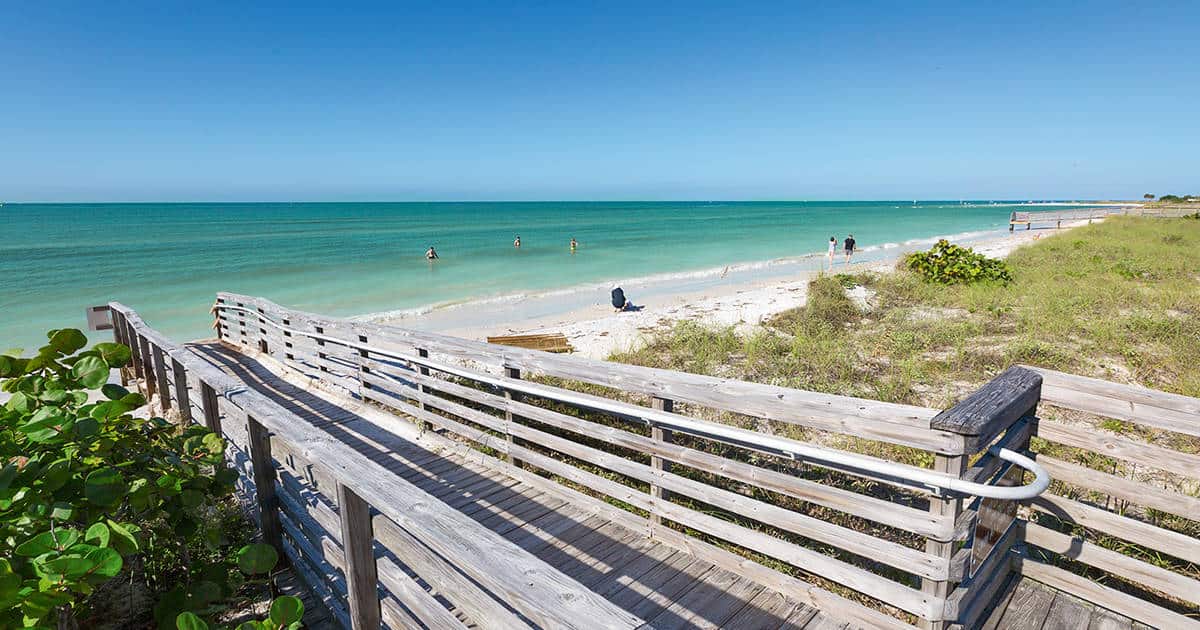 Wooden boardwalk leading to a sandy beach with clear turquoise water under a blue sky. A few people are swimming and relaxing in the water. Green shrubs and grass line the path.