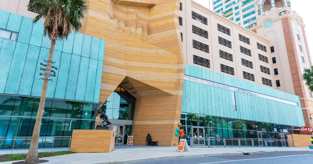 Contemporary building with a unique angular entrance and a mix of tan and teal façades. A sculpture stands near the entrance, and a sandwich board with balloons is positioned on the sidewalk. A tall palm tree is on the left.