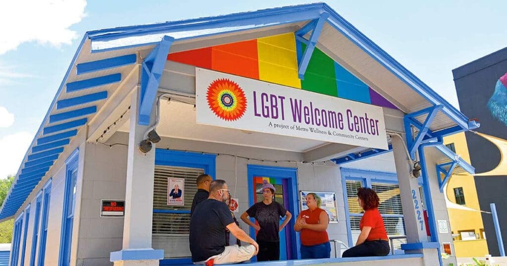 A small, colorful building with rainbow accents and a sign reading LGBT Welcome Center. Several people are chatting on the porch under a bright sky. The structure has blue trim and is adorned with a rainbow stripe near the roof.