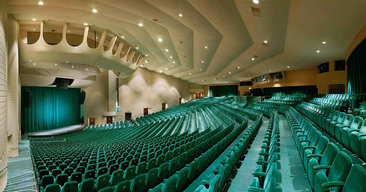 An empty theater auditorium with rows of green seats facing a stage. The walls and ceiling are cream-colored, featuring recessed lighting. Curtains cover the stage, and the spacious hall is well-lit, creating a dramatic and inviting atmosphere.