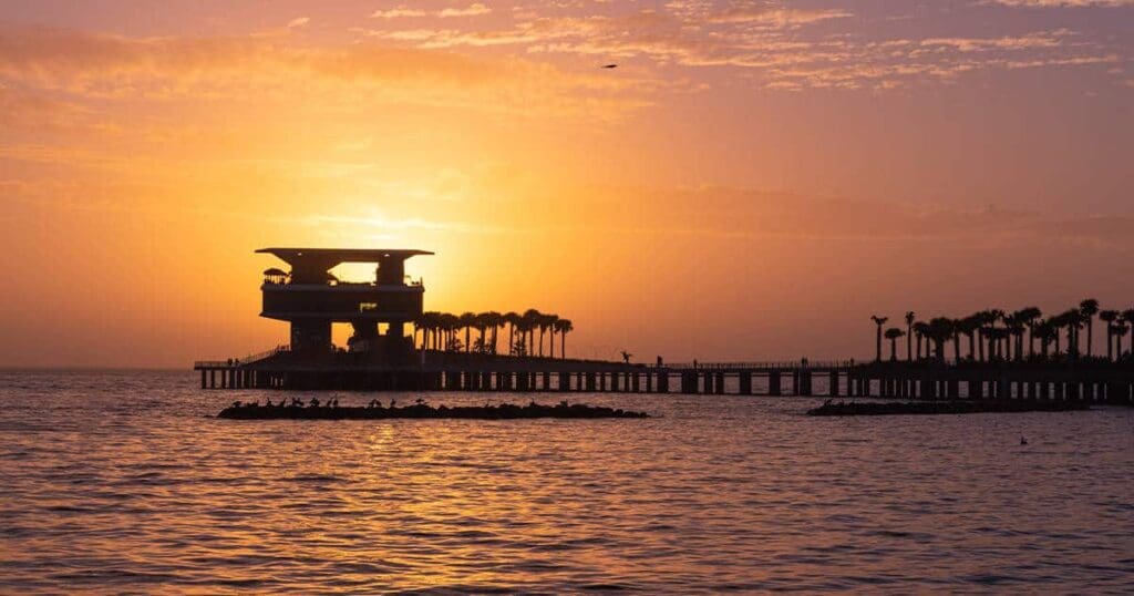 A tranquil sunset over a calm sea with an elevated structure silhouetted against the orange sky. A long pier extends over the water with palm trees lining the walkway, creating a serene coastal scene.