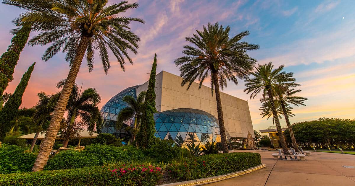 A modern building with a geometric glass facade surrounded by palm trees and lush greenery. The sky is colorful with hues of pink, orange, and blue, suggesting a sunset or sunrise. Benches and walking paths are visible in the foreground.