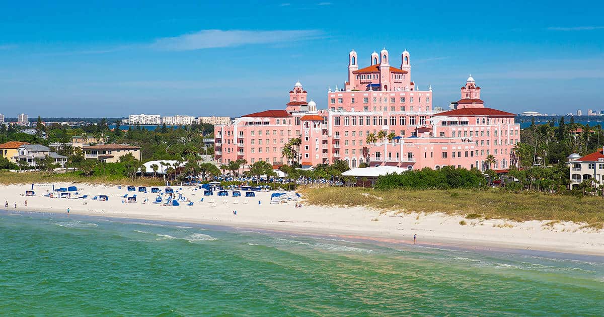 A large pink hotel with red roofing stands majestically on a sandy beach by the turquoise ocean. White cabanas are scattered along the shoreline. The sky is clear and blue, with a distant cityscape visible in the background.