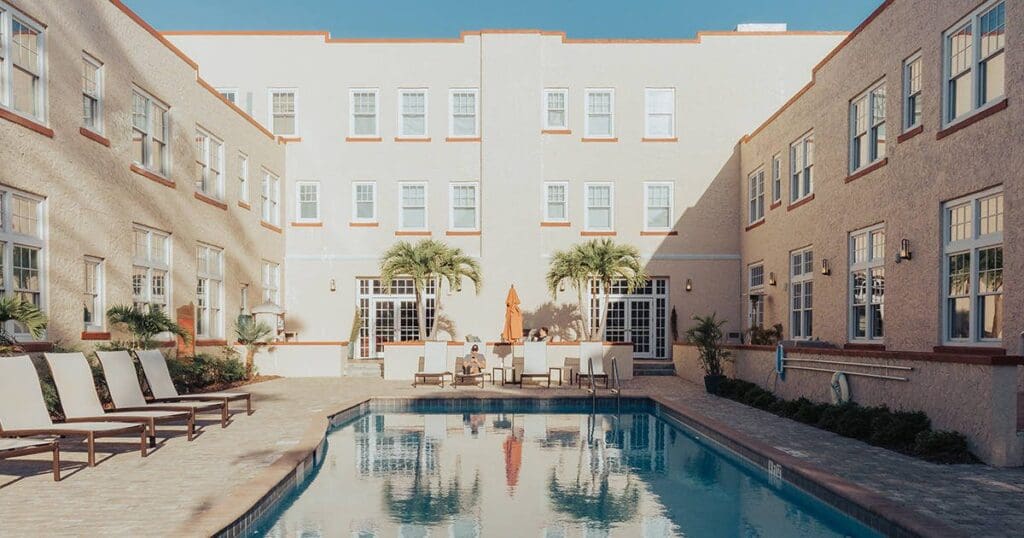 A rectangular swimming pool flanked by beige buildings with multiple windows. Several lounge chairs line the left side, and palm trees decorate the area. A few people sit by a table with an umbrella at the far end. Clear blue sky overhead.