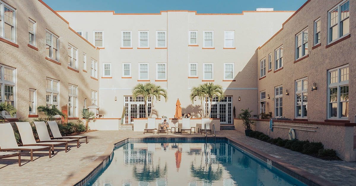 A rectangular swimming pool flanked by beige buildings with multiple windows. Several lounge chairs line the left side, and palm trees decorate the area. A few people sit by a table with an umbrella at the far end. Clear blue sky overhead.