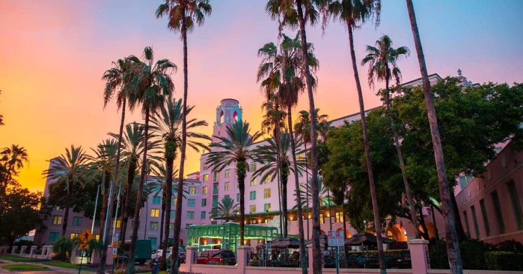 A pink building surrounded by tall palm trees is set against a vibrant sunset sky. The trees create a silhouette effect as they frame the structure, which features arched windows and a circular rooftop section. Lights are visible on the building.