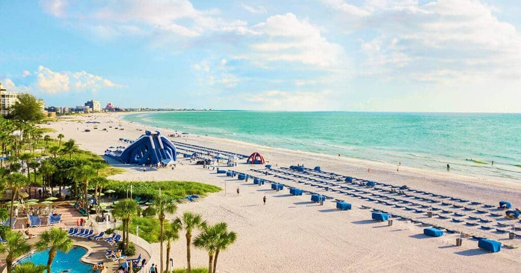 A sunny beach with clear turquoise water and white sand. Blue loungers are lined up on the sand, and inflatable water slides are visible. Palm trees and a few beachgoers are scattered along the shore. Buildings are visible in the distance.