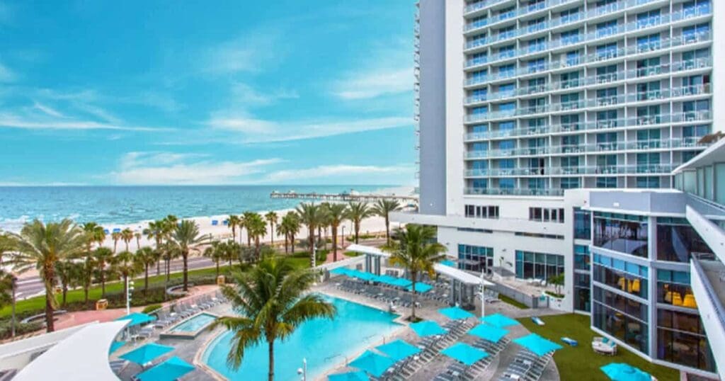 View of a beachfront hotel with a large outdoor pool surrounded by lounge chairs and palm trees. The tall hotel building has numerous balconies. The ocean and sandy beach are visible in the background under a clear blue sky.