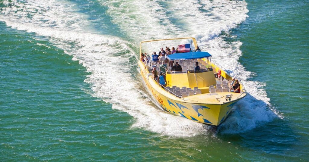 A yellow speedboat with a blue canopy moves swiftly across the green ocean, leaving a trail of white wake. Several passengers are seated, enjoying the ride under a clear, sunny sky. An American flag is visible at the rear.