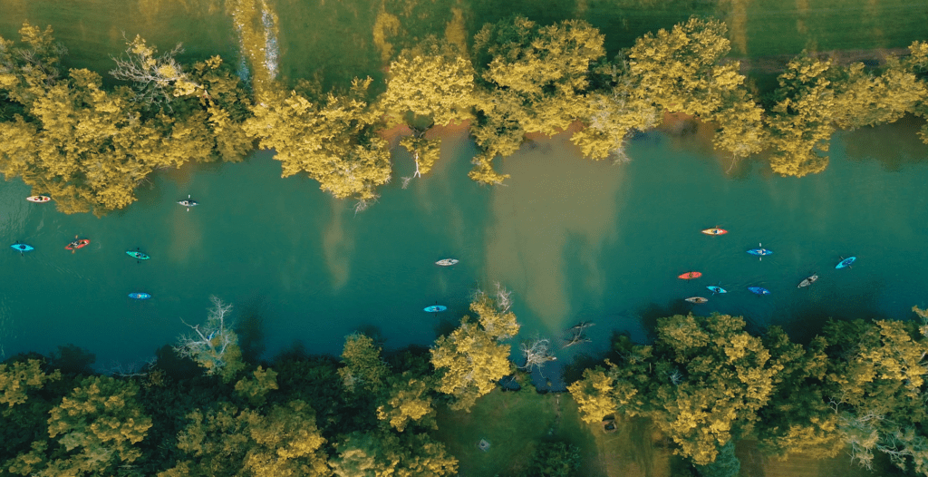 Aerial view of a calm, narrow river surrounded by lush green trees. Colorful kayaks with people paddling are scattered along the water, creating a vibrant contrast against the serene, natural setting.