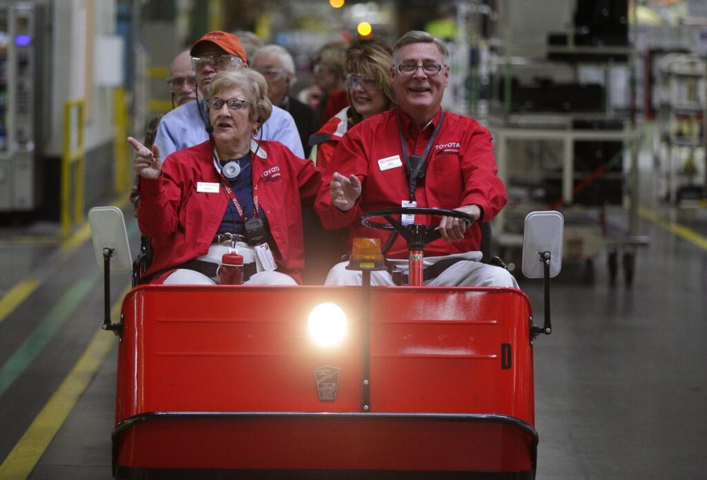 A group of people sit in a red electric cart inside a factory, wearing matching red jackets. They appear to be on a tour, with industrial equipment visible in the background. The front passengers are smiling and engaged in conversation.