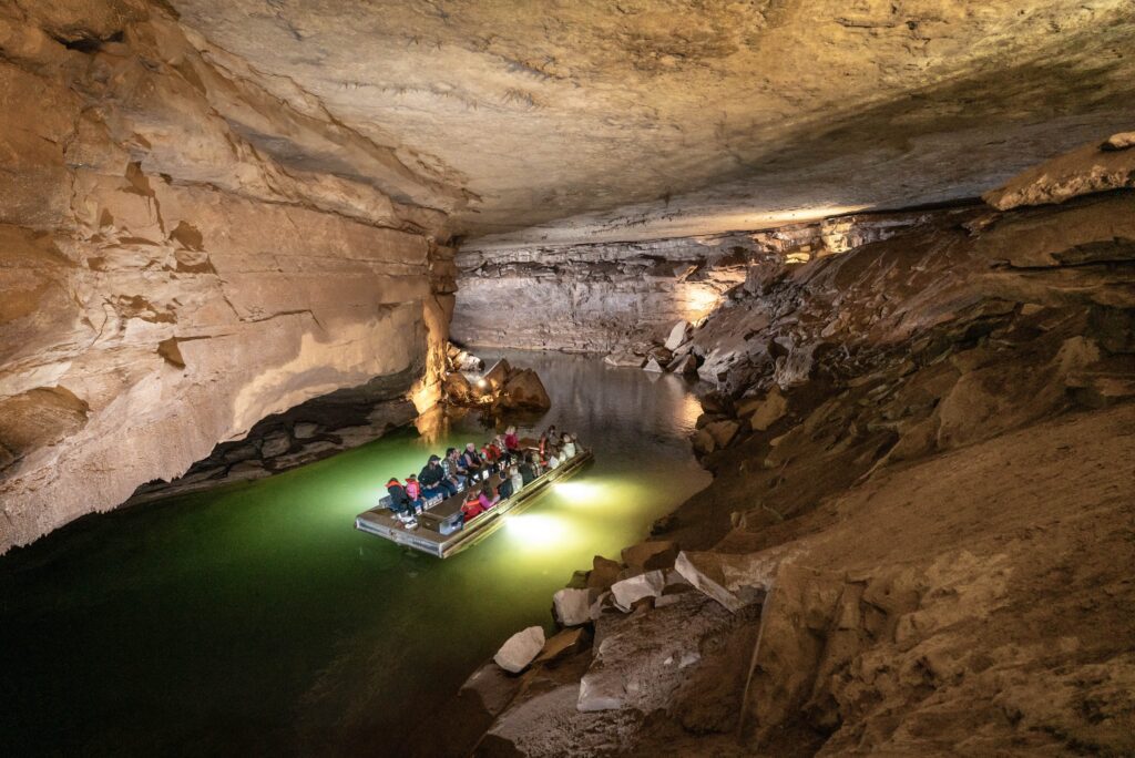 A group of tourists on a small boat explore a large, illuminated cave with rocky walls and a calm greenish water surface. The ceiling is low, and the cave is lit with artificial lights, highlighting its natural rock formations.