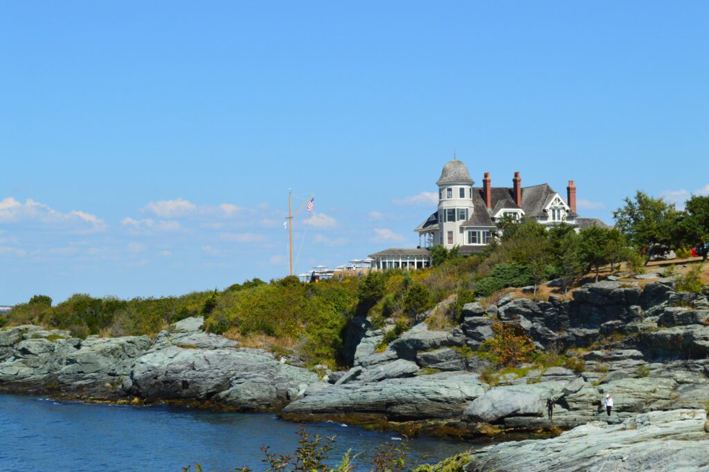 A grand, turreted house sits atop a rocky cliff overlooking the ocean. The sky is clear and blue. A small figure can be seen near the water’s edge. Flags fly on a pole beside the house, surrounded by greenery.
