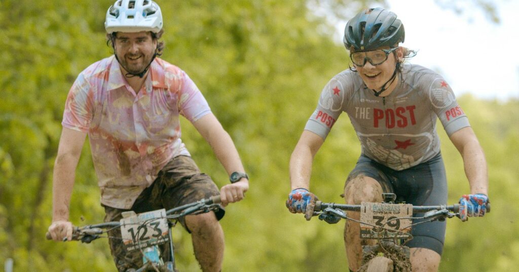 Two cyclists in muddy shirts and helmets ride on a wet, forested trail. The cyclist on the left is wearing a colorful shirt, and the one on the right is in a gray The Post jersey. Both are smiling, surrounded by greenery.