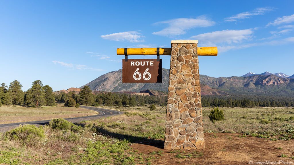 A large stone sign reading Route 66 stands by a winding road. The landscape features green fields and scattered trees, with distant mountains under a clear blue sky.