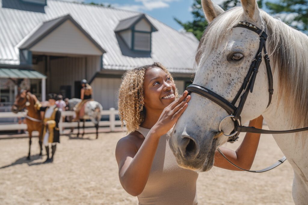 A woman with curly hair smiles while petting a white horse. Behind them, people and horses are visible near a stable with a metal roof. The scene is bright and sunny.