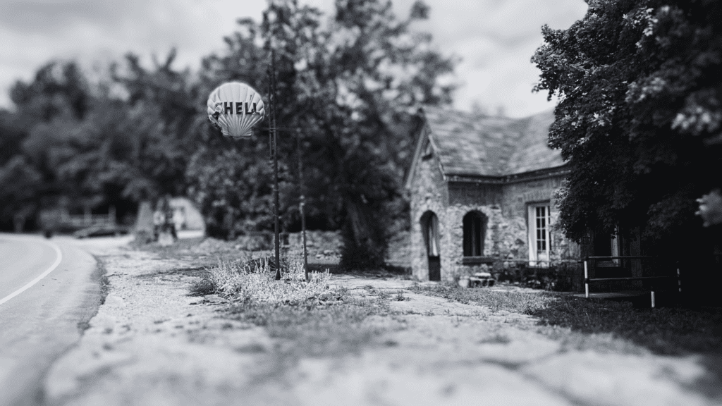 A black and white image of a vintage Shell gas station. An old brick building sits beside a road, with a round Shell sign on a pole. The area is overgrown with grass and trees, giving a nostalgic feel.