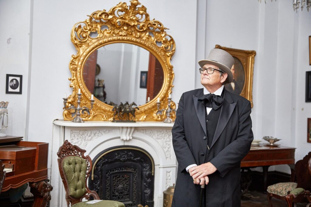 A person in a top hat and formal historical attire stands in a vintage room. The room features an ornate gold-framed mirror, a white fireplace, and antique furniture, creating a 19th-century ambiance.