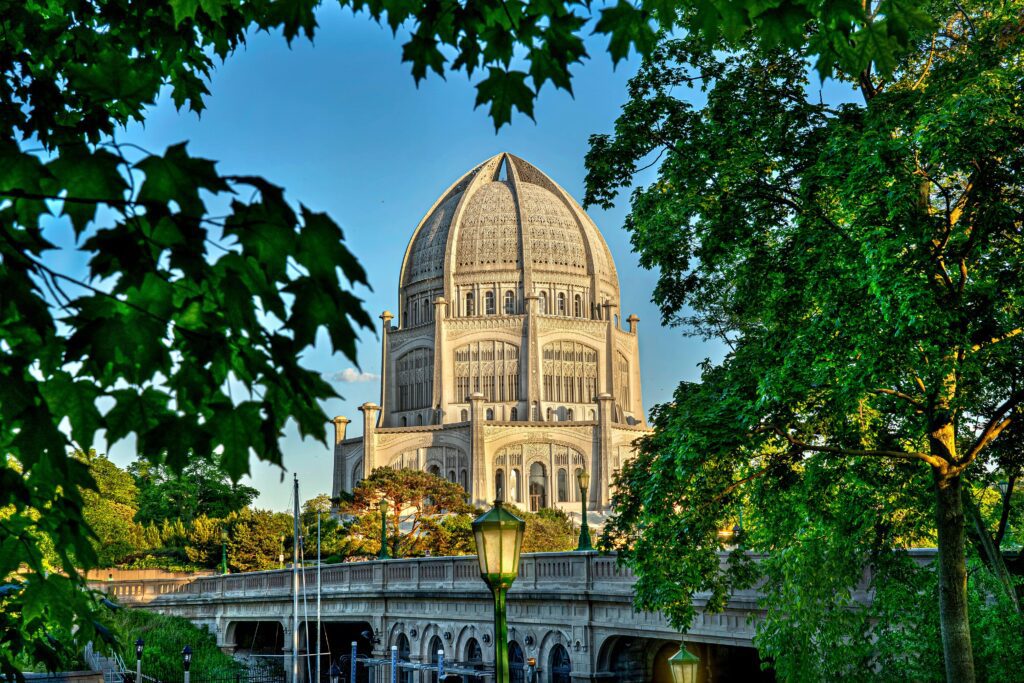 The image shows a large, ornate building with a domed roof, surrounded by lush green trees. A stone bridge with arches is in the foreground, and a lamp post is visible on the path leading to the building.