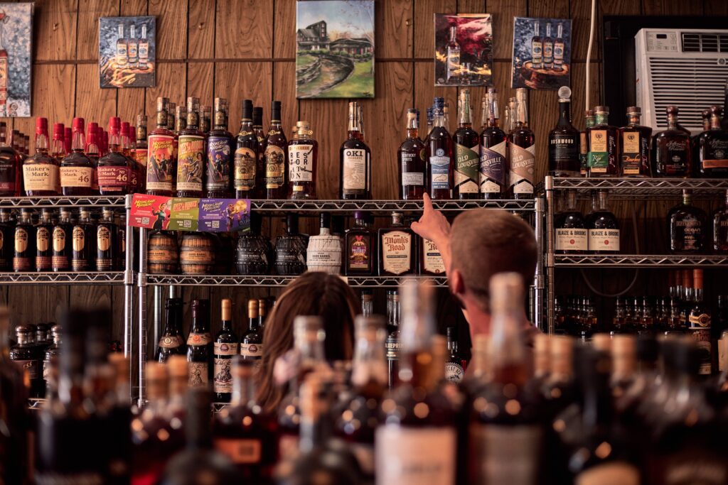 Two people browse liquor bottles on metal shelves in a store. One person points at a bottle. The shelves are filled with various spirits, and the wooden wall behind features small paintings and an air conditioner.