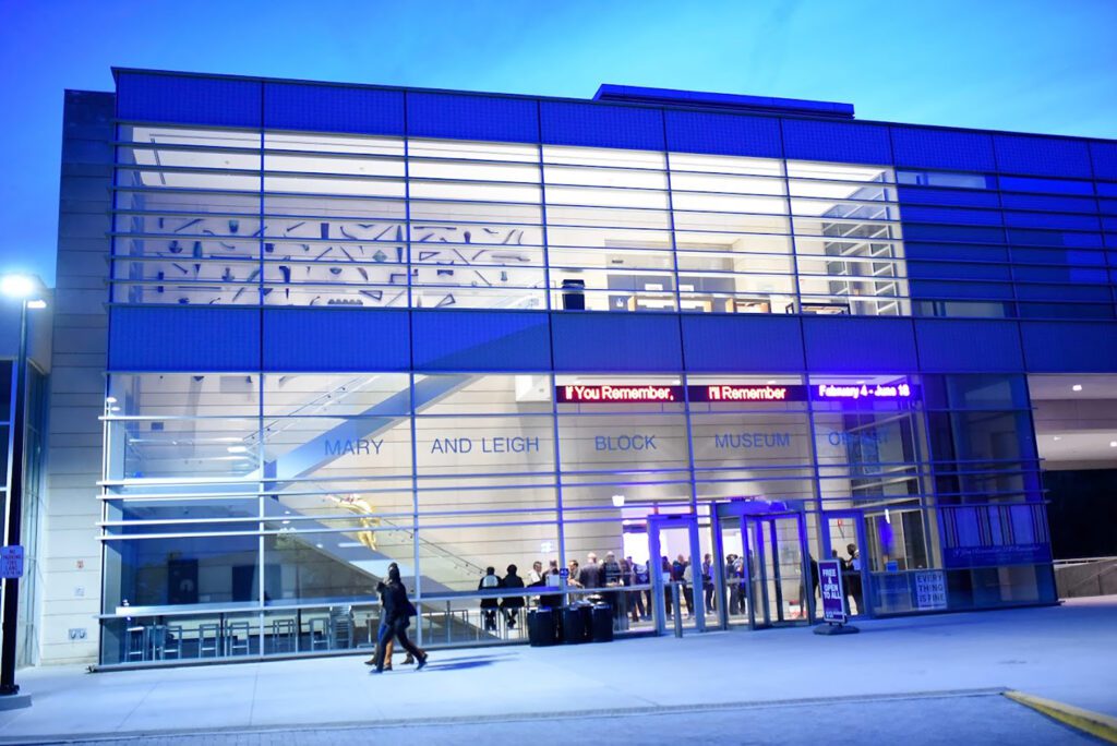 Modern building facade with glass walls and a prominent entrance. People are gathered near the entrance under signage that includes the words I You Remember. The scene is illuminated by blue and white lights in the evening.