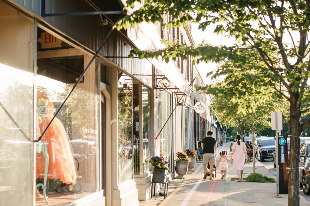 A family walks down a sunlit sidewalk in front of a row of shops. Storefronts display various items, including a display with an orange dress. Trees line the street, casting dappled shadows on the pavement.