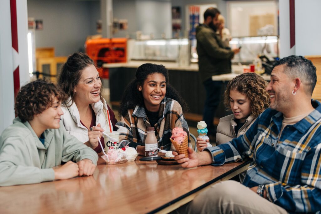 A family of five sits at a wooden table enjoying ice cream. They are smiling and engaged in conversation. The setting appears to be an ice cream parlor, with display cases and customers visible in the background.