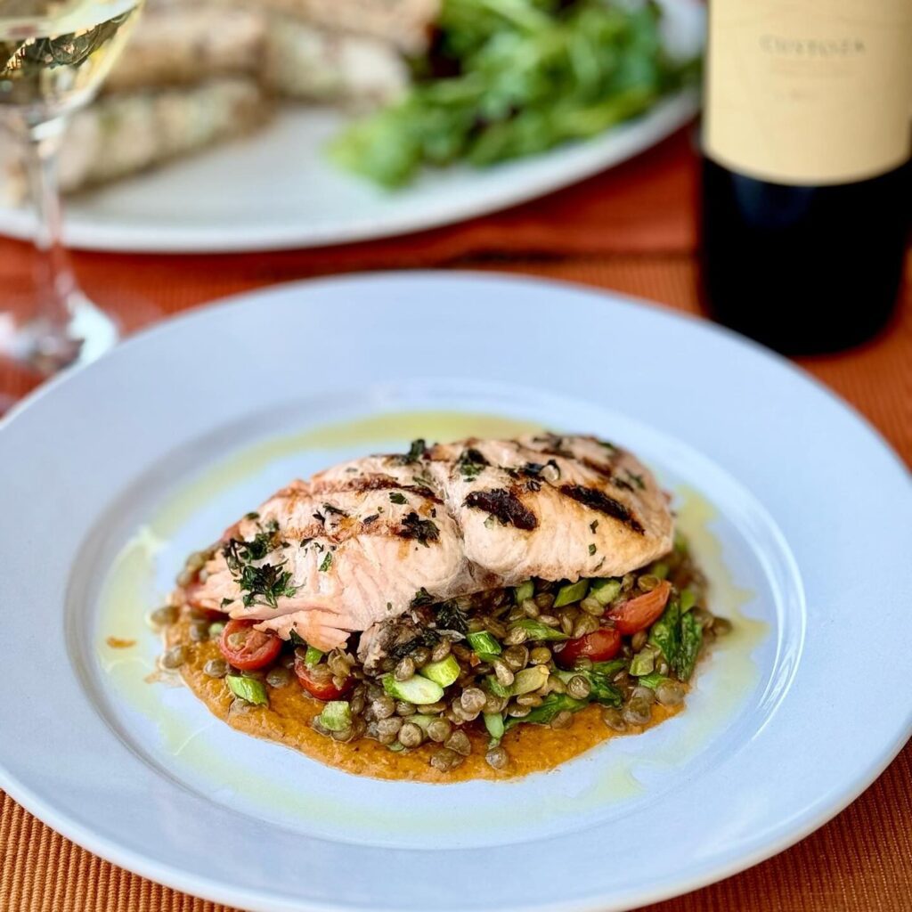 Grilled salmon fillet topped with herbs on a bed of lentils, asparagus, and cherry tomatoes, drizzled with sauce on a white plate. A glass and bottle of wine, along with a salad, are visible in the background.