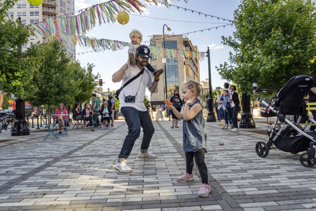 A man with a child on his shoulders dances with a little girl in an outdoor plaza decorated with colorful streamers. People are seated in the background, and a stroller is on the right. The atmosphere is lively and festive.