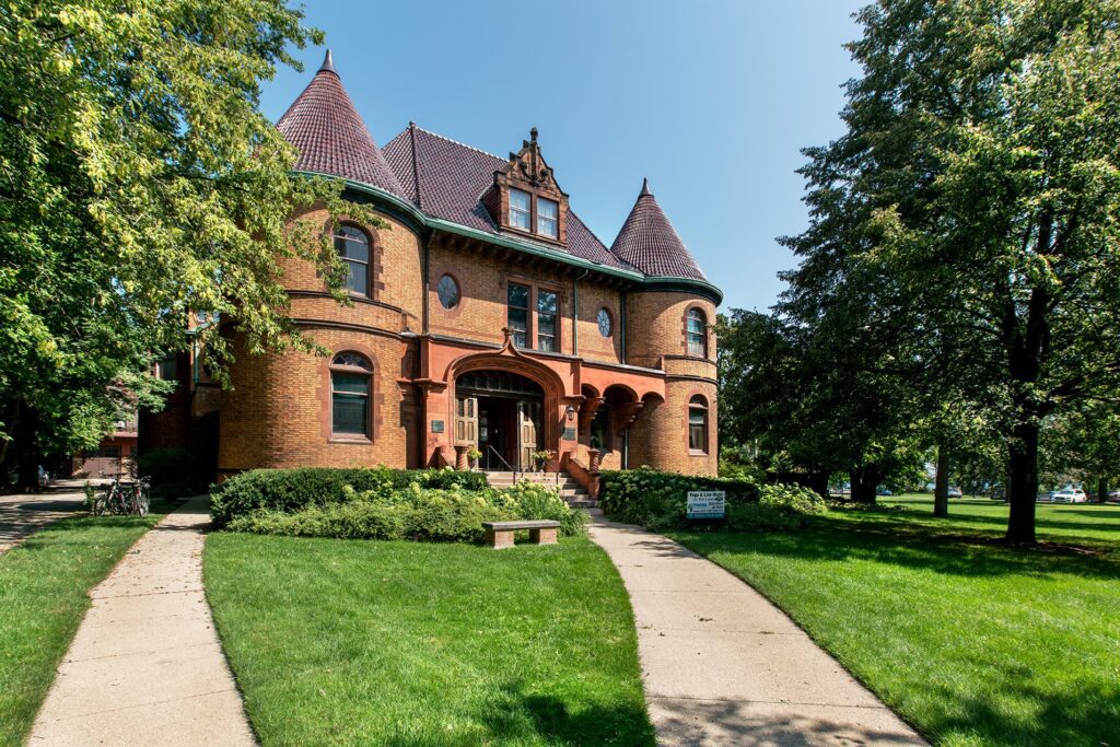 A large, historic brick mansion with two round towers, steep roofs, and arched windows sits amidst a lush lawn. Two concrete paths curve toward the entrance. Trees frame the scene under a clear blue sky.