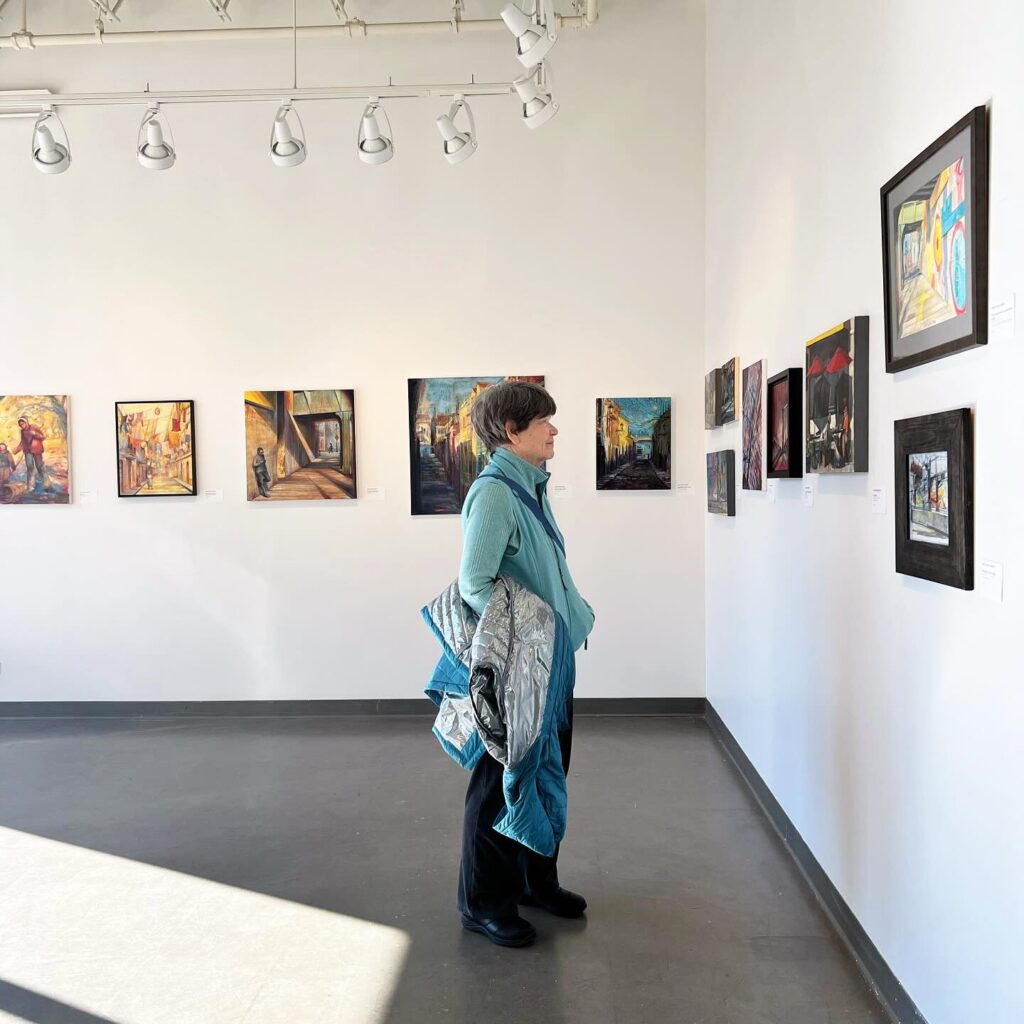 Person in a light blue top and black pants stands in an art gallery, observing framed artworks on white walls. Natural light pours in from a window, casting shadows on the floor. Track lighting illuminates the paintings.