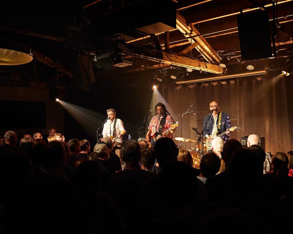 A band performs on a dimly lit stage with three guitarists/singers under warm spotlights. The audience is closely gathered, enjoying the show. The stage has a rustic, exposed ceiling with wooden beams.