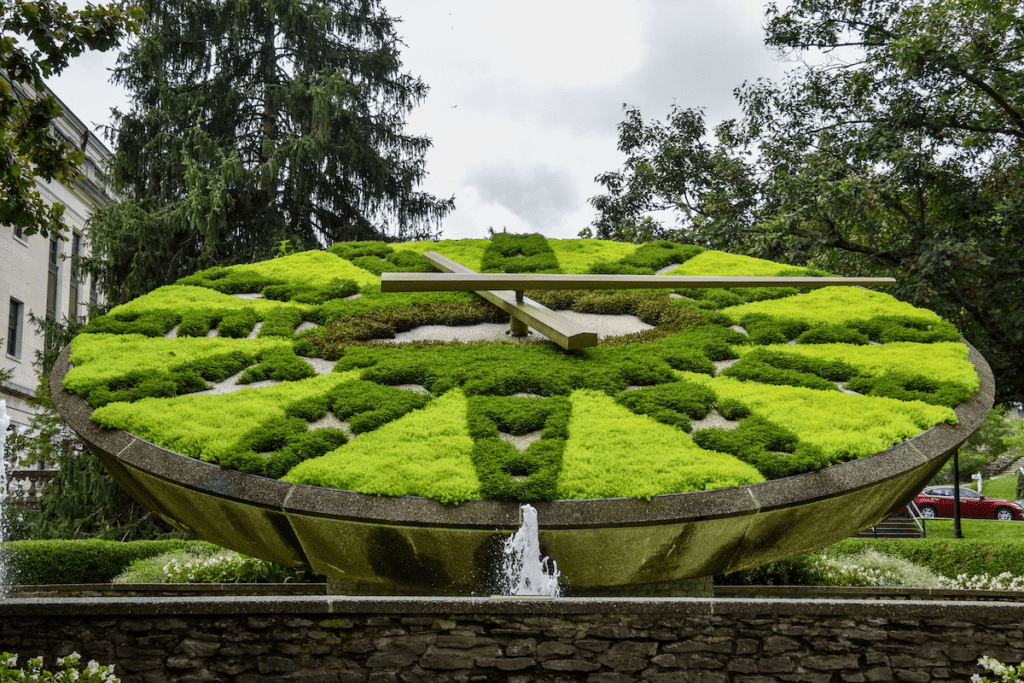 A large outdoor floral clock with lush green and yellow plants forming the clock face and numbers. Two wooden clock hands indicate the time. Trees and a building are visible in the background.