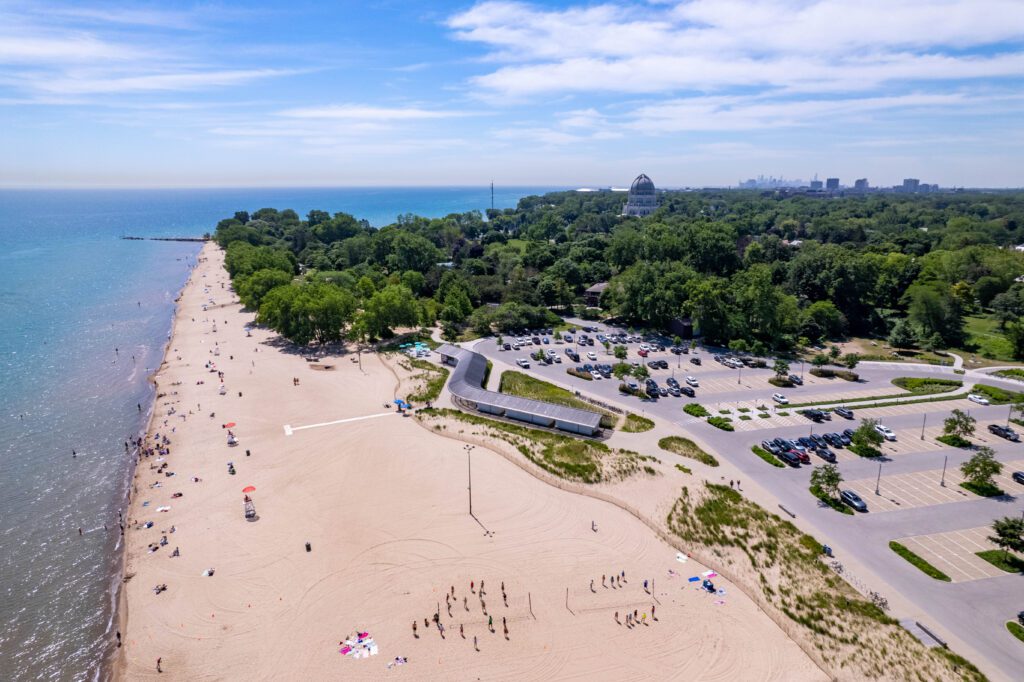 Aerial view of a beach with scattered sunbathers near the water. A parking lot is adjacent to the sandy shore, met by a lush green forest. A few sports games are happening in the sand. A blue sky with light clouds stretches above.