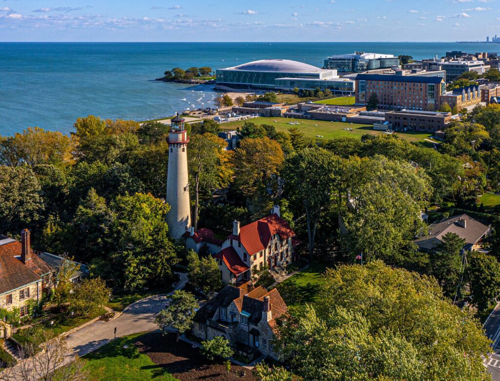 Aerial view of a coastal area with a tall lighthouse surrounded by trees and historic buildings. In the background, a large sports arena and modern buildings are near a lake. The scene is under a clear blue sky.