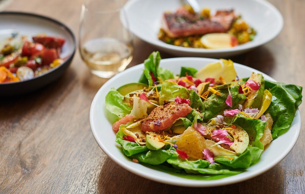 A white bowl filled with a fresh salad featuring lettuce, grapefruit slices, potatoes, and garnished with colorful petals. In the background, there are two blurred plates of food and a glass on a wooden table.