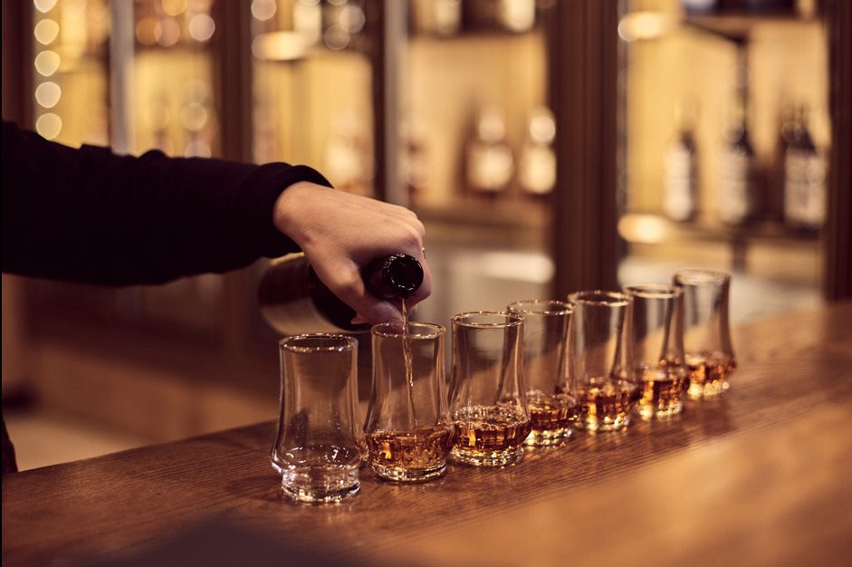 A person pours whiskey from a bottle into several tulip-shaped glasses lined up on a wooden bar counter. Shelves with bottles are visible in the blurred background, creating a warm, inviting atmosphere.