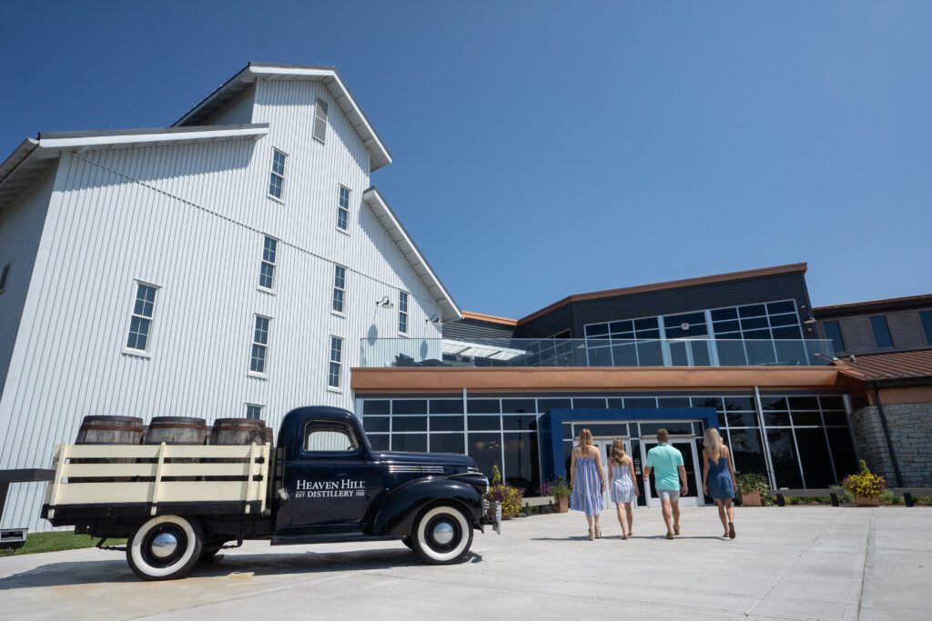 A group of four people walk toward a large building with modern and rustic architecture. On the left, an old truck displays barrels on its bed, with Heaven Hill written on the side. The sky is clear and blue.