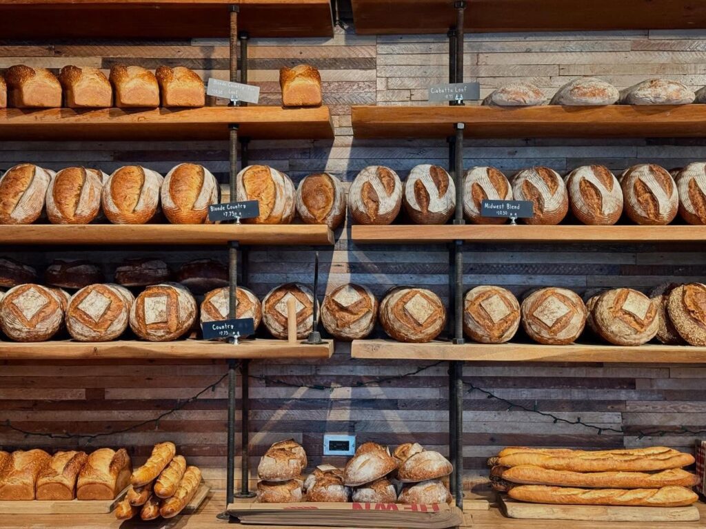 A bakery shelf displays various types of bread, including round loaves with decorative scoring and baguettes. They are arranged neatly on wooden shelves against a brick-style wall. Small signs label different types of bread.